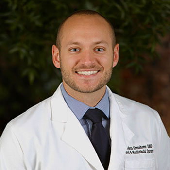 The image shows a smiling man wearing a white lab coat, standing outdoors with a tree behind him, posing for the photograph.