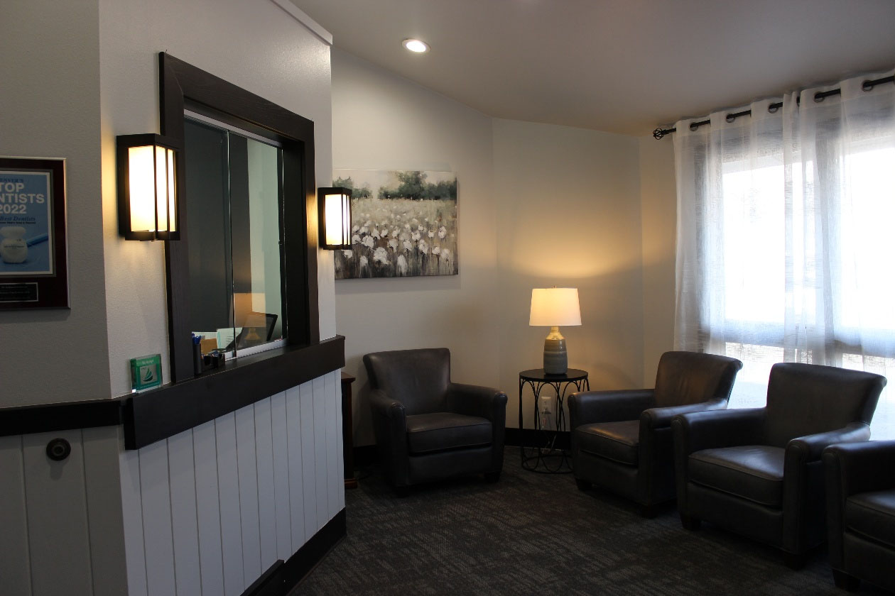 The image depicts a waiting area with modern furniture, including chairs and a couch, arranged around a central table with a lamp on it. A large window with curtains allows natural light into the room.