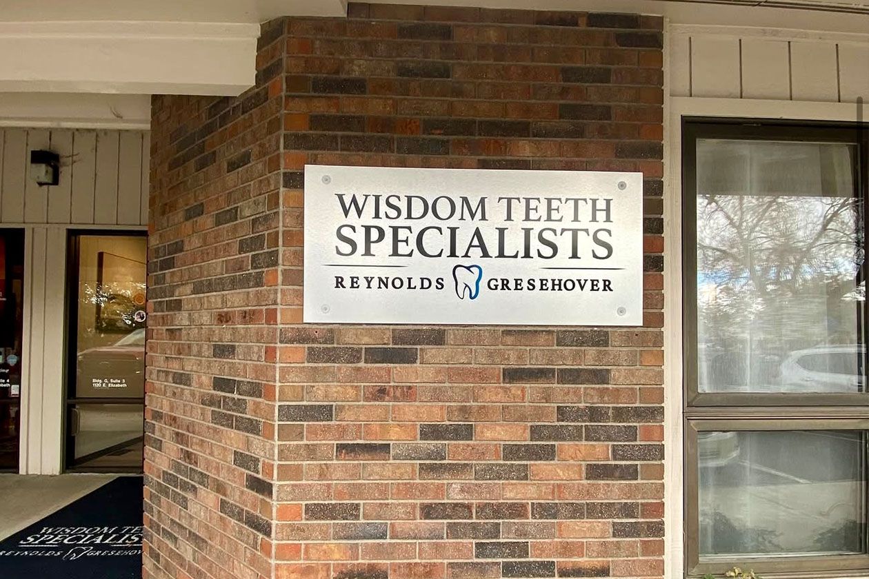 The image shows a sign with the text 'WISDOM TEETH SPECIALISTS' and below it, the name 'RJ Reynolds'. The sign is mounted on a brick wall outside a building, which has a red and white color scheme.