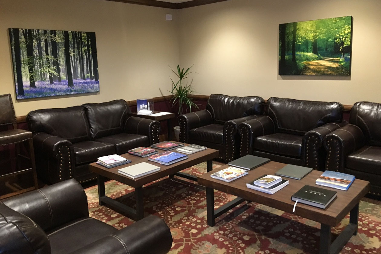 The image shows a spacious waiting area with multiple brown leather sofas arranged around a central coffee table. On the wall, there s a large framed painting of a forest scene.