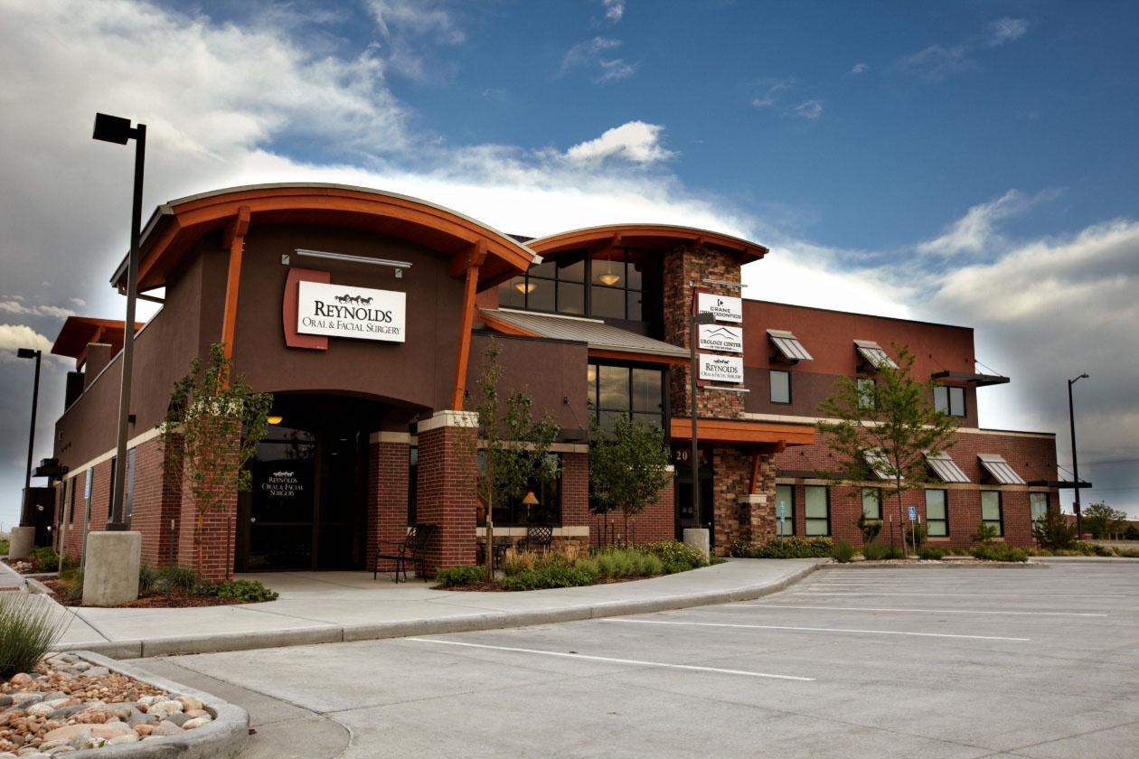 The image shows a two-story building with a prominent sign that reads  Rocky Mountain Lodge.  It appears to be a hotel or motel, given its architectural style and the presence of multiple doors and windows. The exterior walls are painted in shades of brown and tan, and there is a covered entrance area with lighting fixtures. The building is situated on a paved parking lot, and the sky above is partly cloudy.