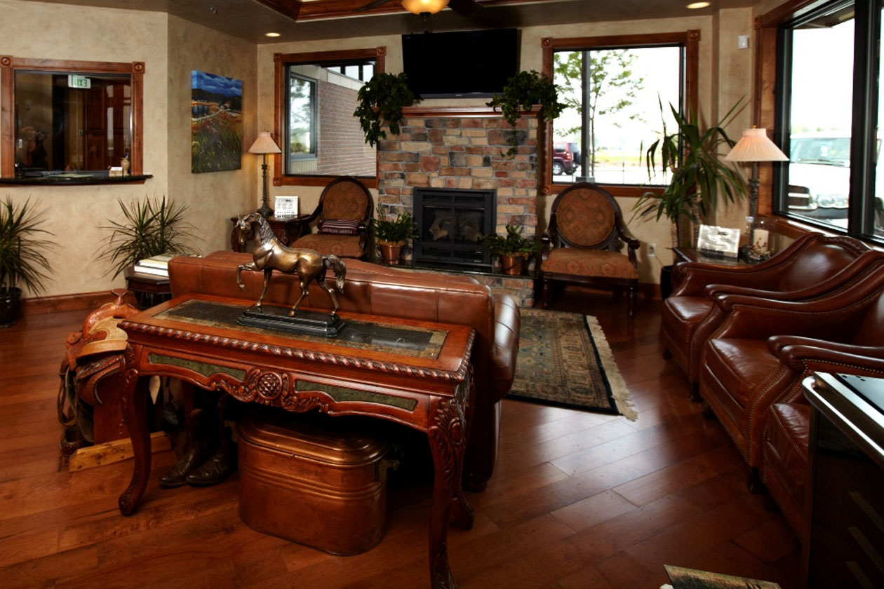 The image shows an interior view of a spacious living room with hardwood floors, a stone fireplace, leather furniture, and various decorative elements such as framed pictures on the wall and potted plants.