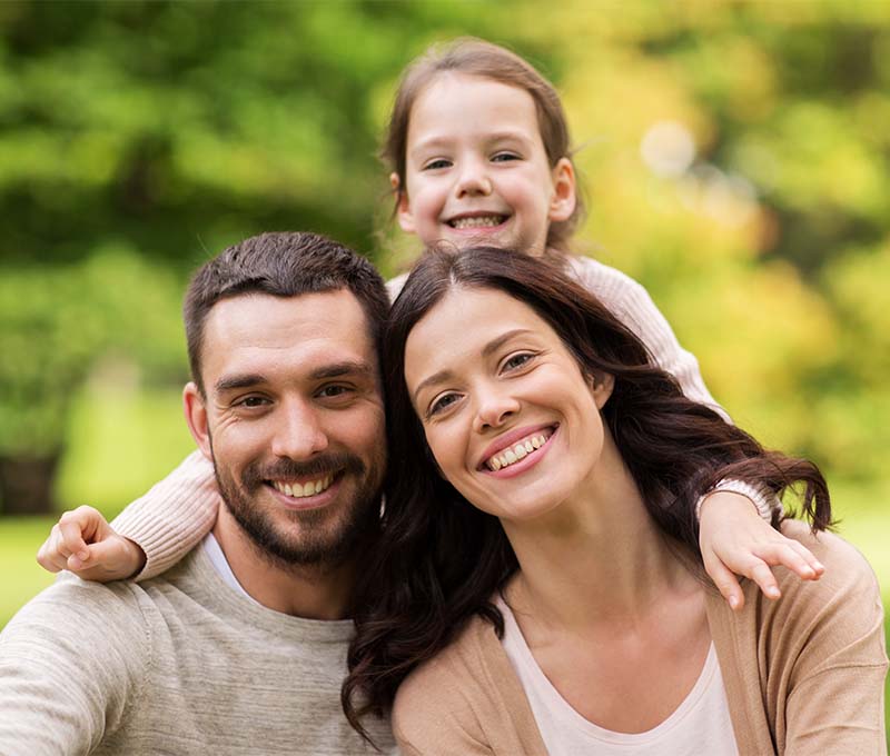 This is a color photograph featuring a family with three individuals  an adult male, an adult female, and a young child. They appear to be outdoors during daylight, posing for the photo with smiles on their faces. The adults are embracing each other, and the child is in front of them, looking at the camera.