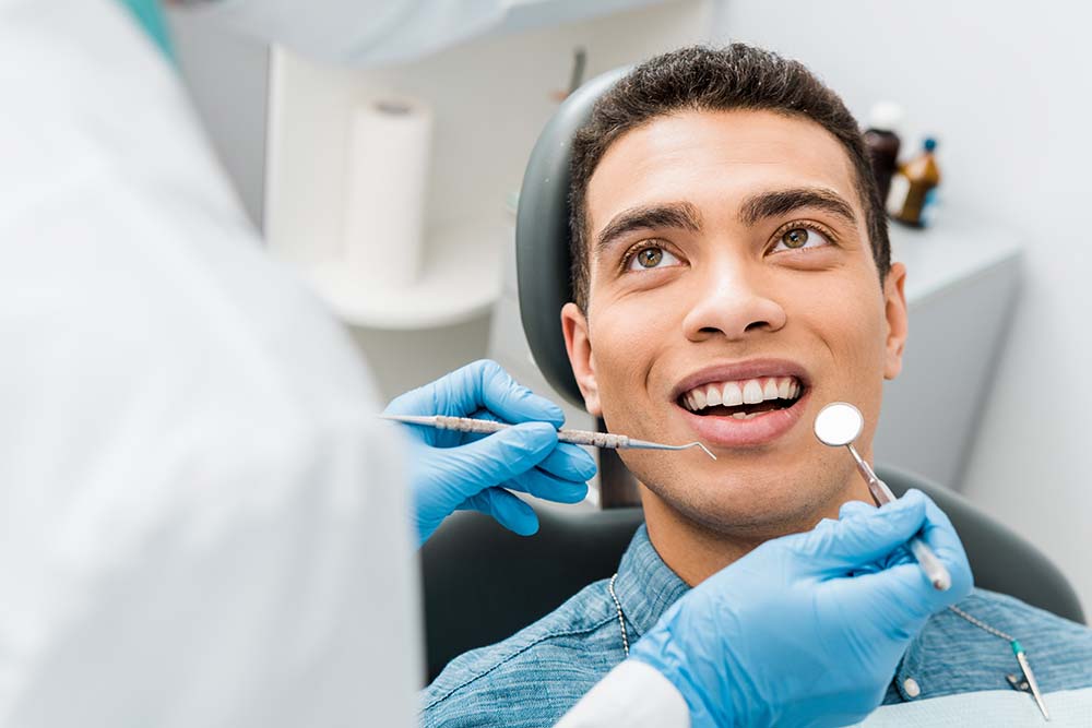 The image shows a dental professional performing a procedure on a patient s teeth, with the patient smiling and looking up at the camera, both wearing protective eyewear and gloves.