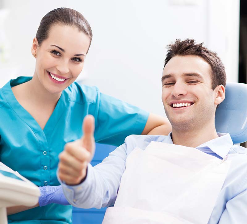 A smiling dental patient giving a thumbs-up sign while seated in a dental chair with a dental professional standing beside him, both in a dental office setting.